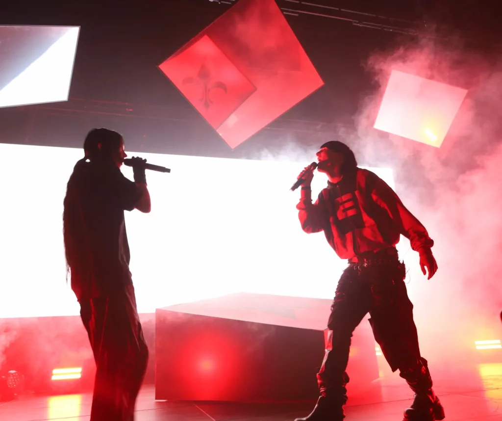 Deux artistes chantent face à face sur une scène rouge enfumée, avec des écrans lumineux suspendus au-dessus d’eux.