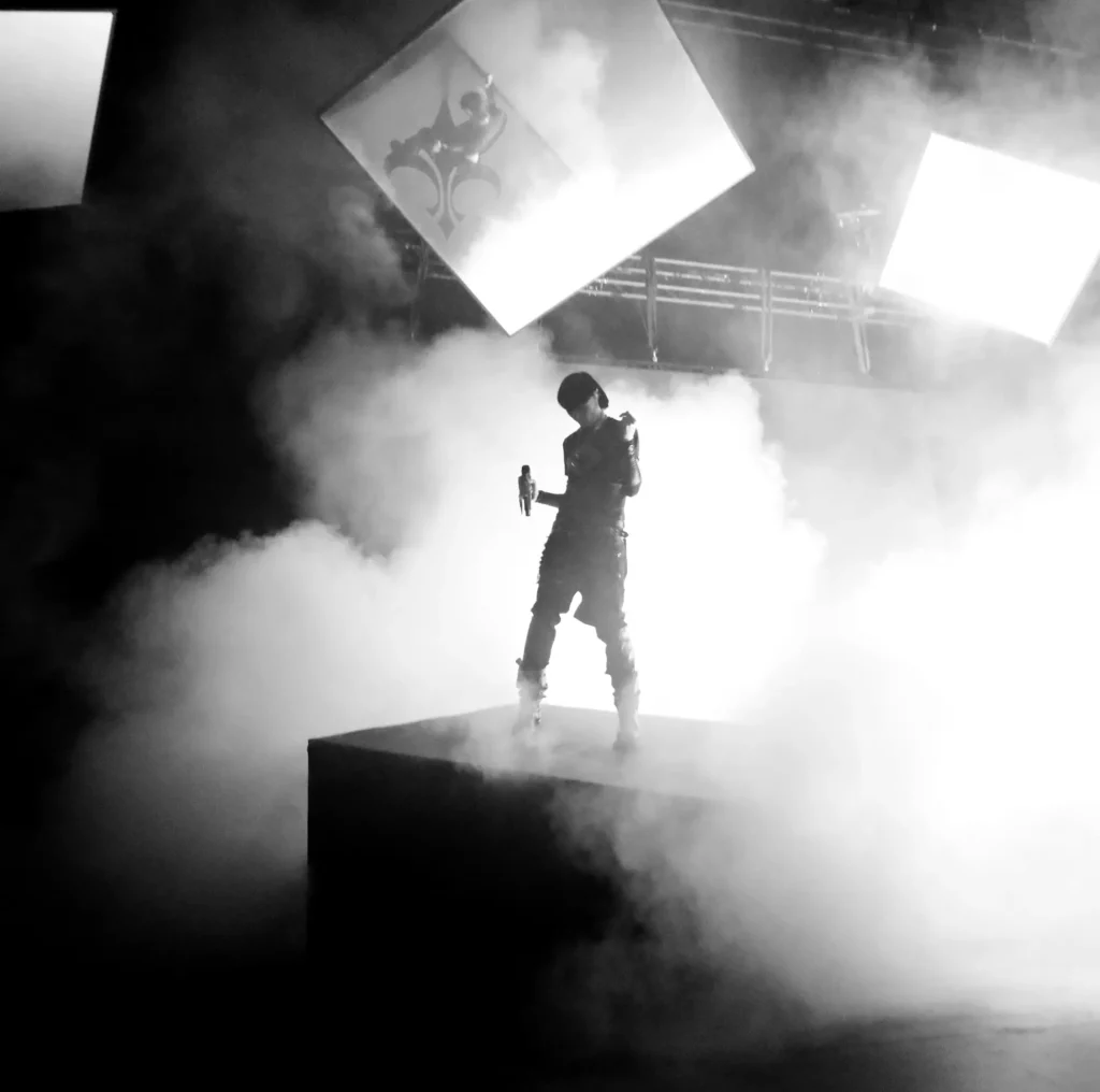 Artiste debout au centre d’une scène envahie de fumée, éclairée par des écrans lumineux suspendus.