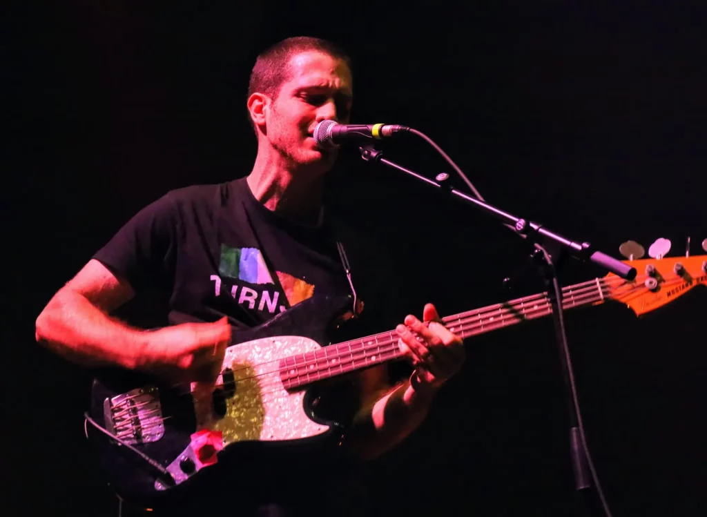 Musicien jouant de la basse et chantant au micro sur une scène au fond noir.