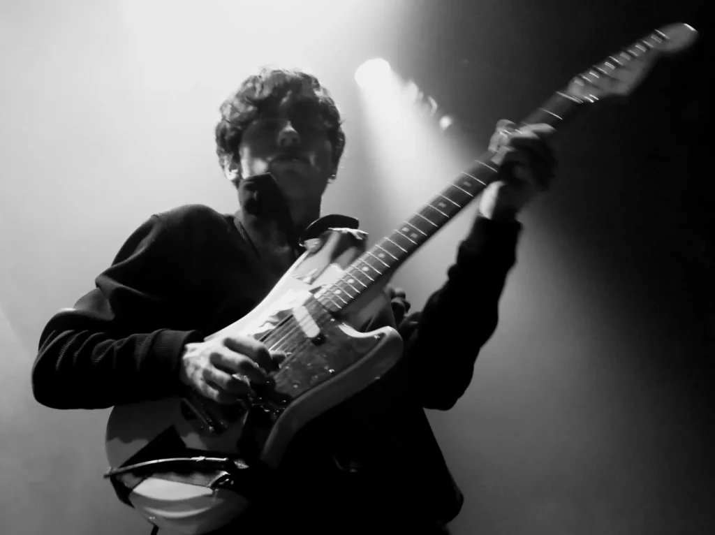 Guitariste jouant sur scène dans une lumière tamisée, capté en noir et blanc.