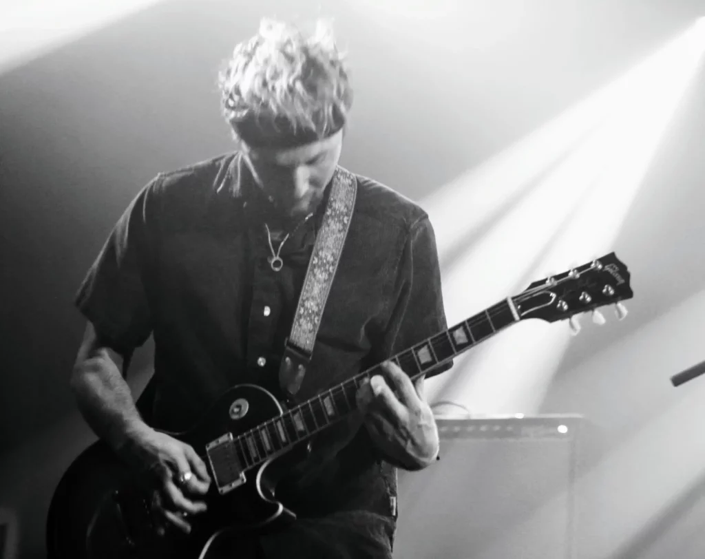 Image en noir et blanc avec un guitariste jouant sur scène, éclairé par des faisceaux lumineux.