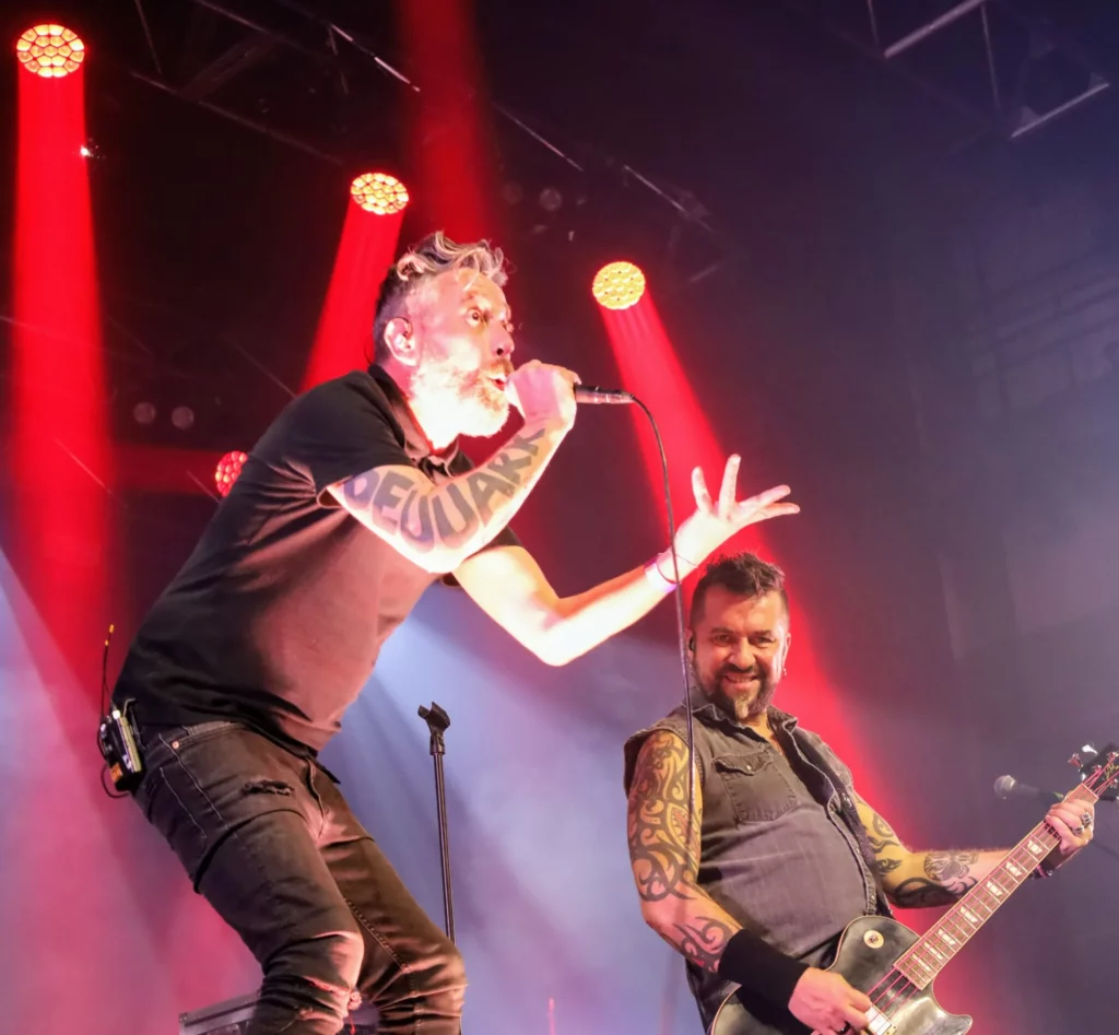 Le chanteur et le guitariste des Fatals Picards sur scène pendant un concert, sous des lumières de scène colorées en rouge et blanc.