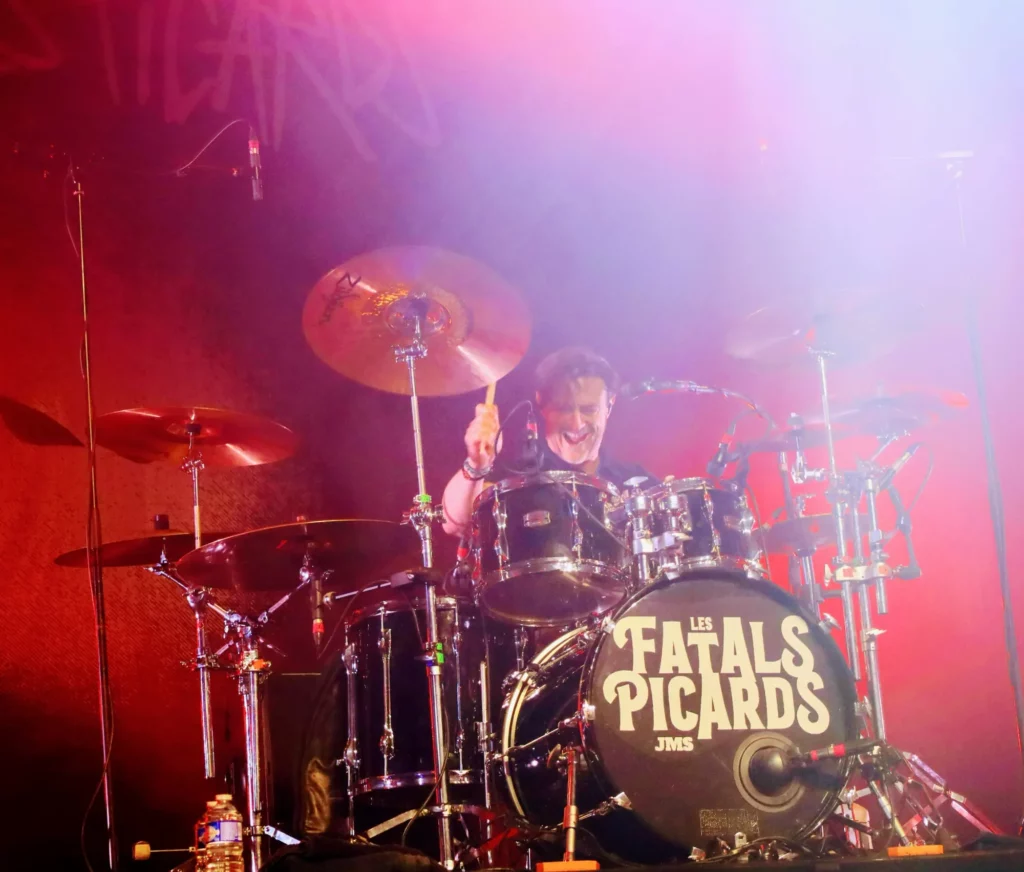 Le batteur des Fatals Picards en concert sur scène, jouant de la batterie sous un éclairage rouge avec l'inscription du nom du groupe sur la batterie.