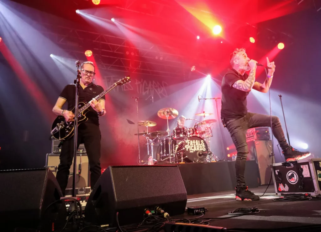 Les Fatals Picards en live sur scène lors d’un concert, avec chanteur, guitariste et batteur illuminés par des spots rouges et blanc.