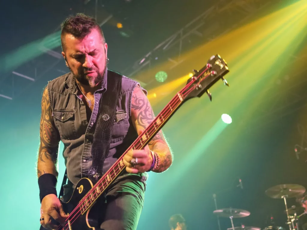 Le guitariste des Fatals Picards au premier plan sur scène, illuminé par des spots verts et orange, avec le batteur en arrière-plan pendant le concert.