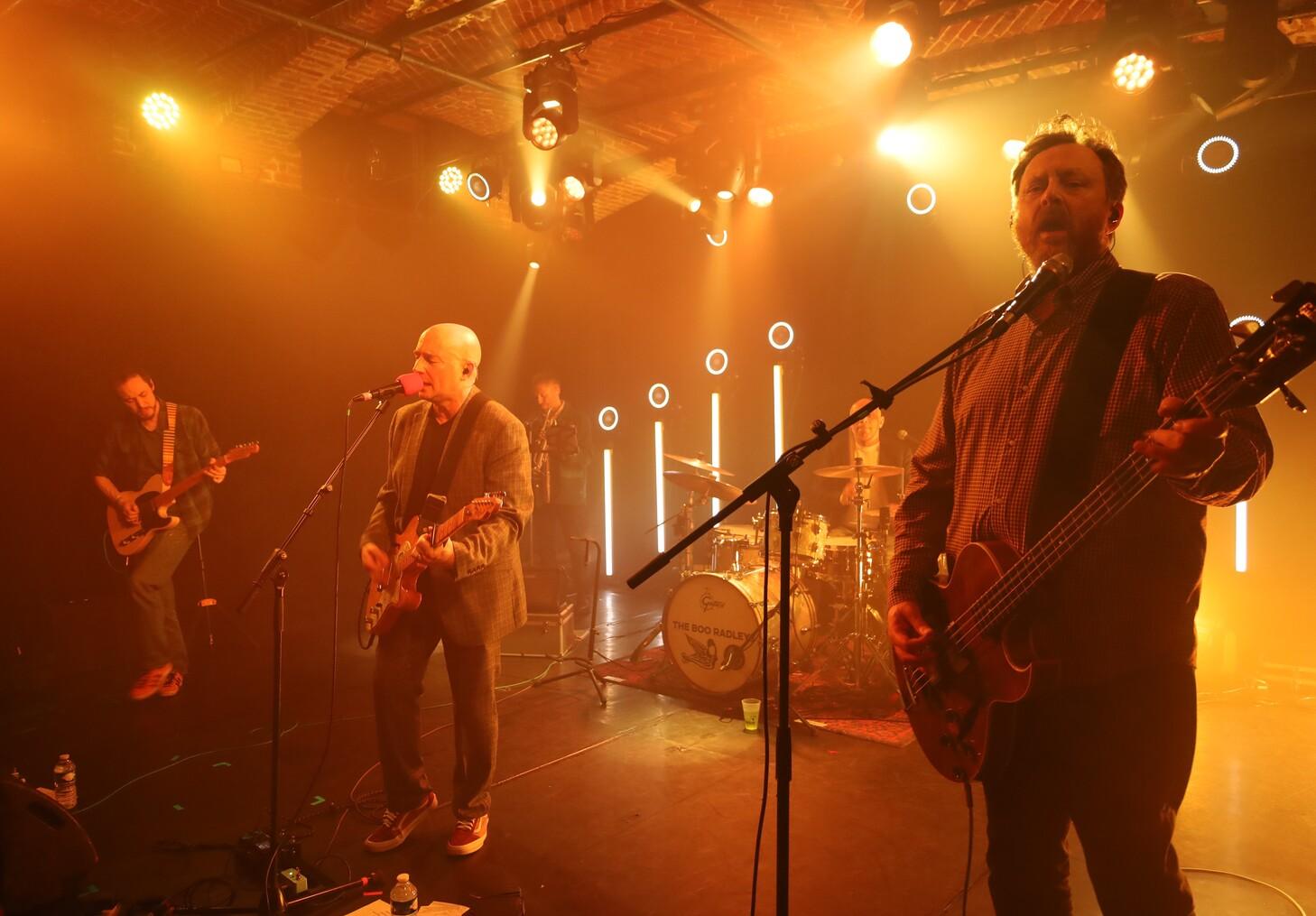 Groupe de rock sur scène jaune-orangée : chanteur barbu au micro central, guitariste chauve à gauche, bassiste barbu à droite jouant basse, batterie, éclairages spots ronds et bâtons lumineux, ambiance live chaleureuse.