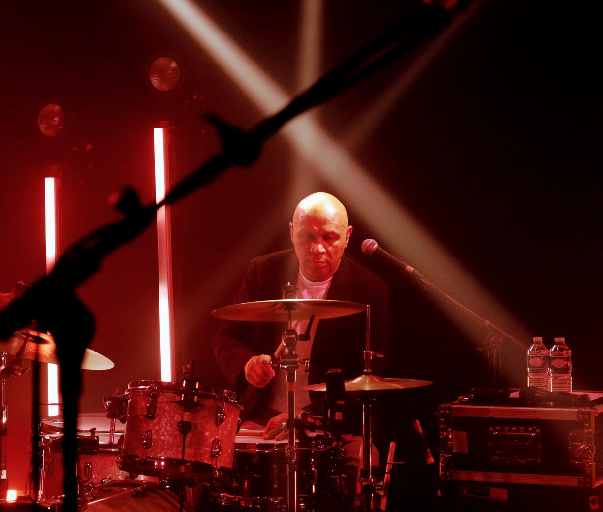 Batteur chauve concentré derrière sa batterie rouge/noire, cymbales et baguettes en action, éclairages rouges et blancs croisés, bouteilles d’eau à côté, ambiance concert live sombre.