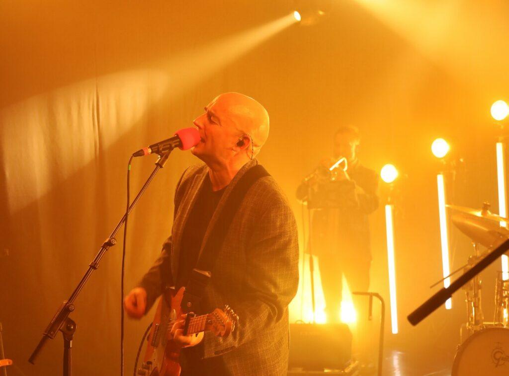 Chanteur-guitariste chauve au micro rose en bouche, guitare en main, veste grise, éclairages orange-jaune et bâtons lumineux, musicien flou (trompettiste ?) en fond, scène live chaleureuse.