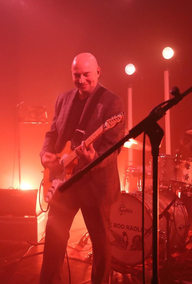 Guitariste chauve souriant du groupe Boo Radleys en veste grise, jouant guitare électrique sur scène rouge-orangée, batterie Gretsch en arrière-plan, éclairages spots blancs, concert live.