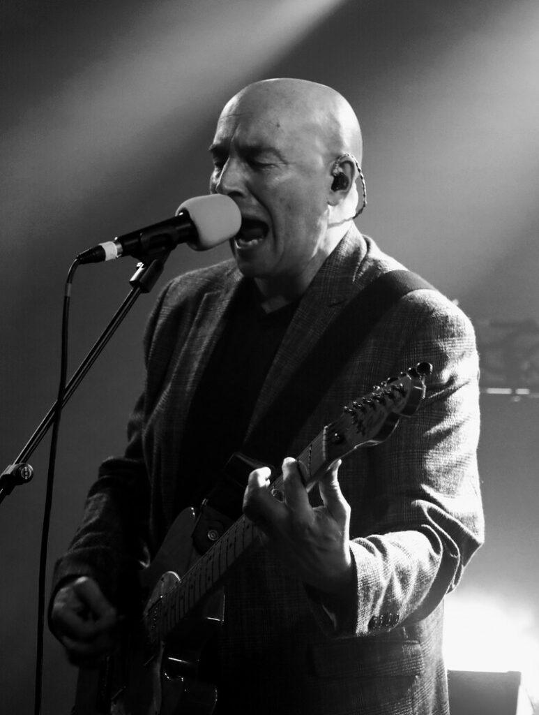 Portrait en noir et blanc d’un chanteur-guitariste chauve en veste grise, micro blanc à la bouche, expression passionnée, guitare électrique, éclairages scène live contrastés.