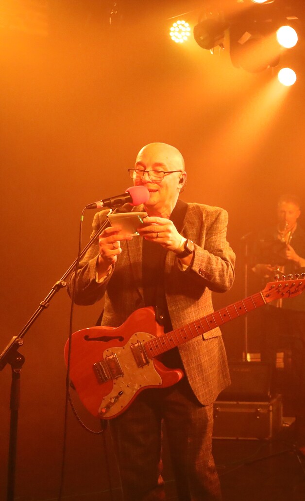Guitariste chauve à lunettes souriant, micro rose en bouche et smartphone en main, guitare orange semi-acoustique, veste grise, scène orange illuminée par spots, musicien flou en fond live.