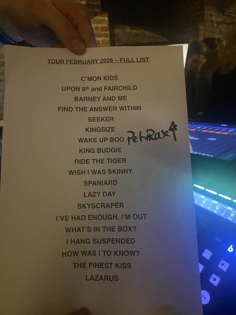 Feuille de setlist de concert ‘TOUR FÉVRIER 2025 – FULL LIST’ tenue en main devant une console de mixage éclairée en bleu-violet, listant chansons comme ‘Baroness and the Fairchild’, ‘Wake Up Boo’, ‘Pet Rock’ (signature manuscrite), ‘Ride the Tiger’, ‘Lazarus’, sur fond sombre avec effets lumineux.