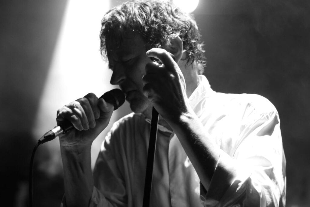 Chanteur live homme cheveux bouclés chemise blanche micro geste main scène concert noir et blanc éclairage spotlight