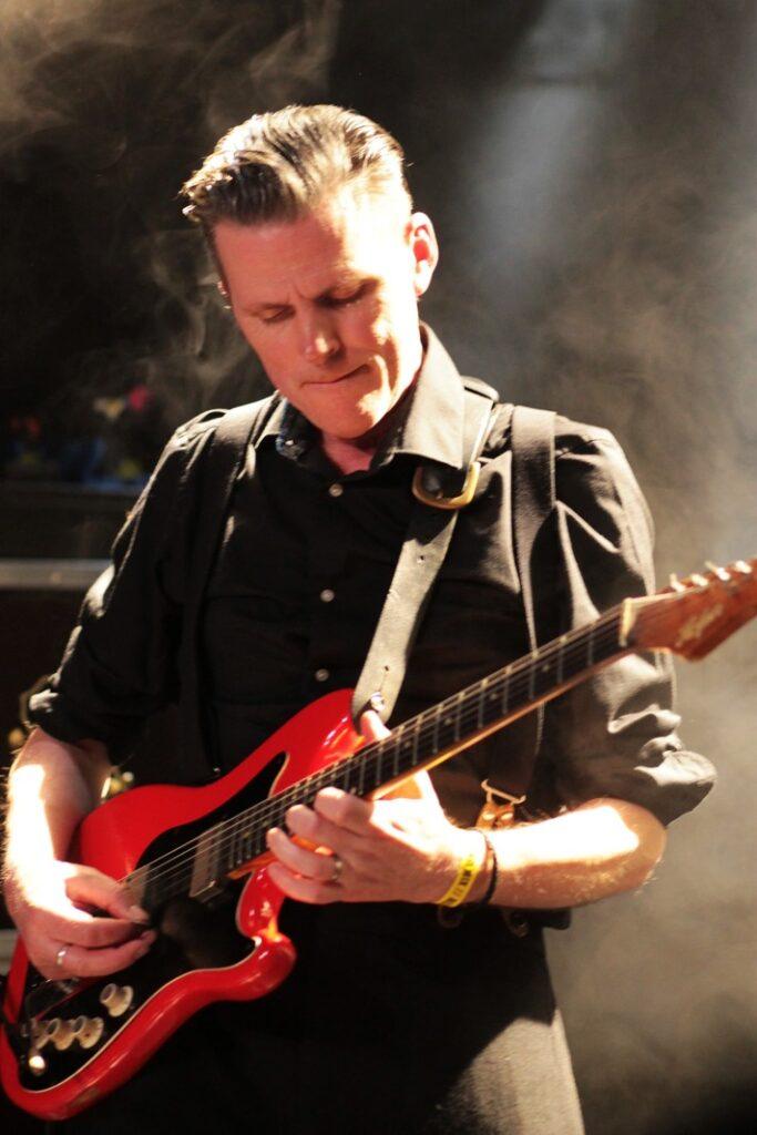 Guitariste rock sur scène brumeuse, cheveux gominés, chemise noire bretelles cuir jouant guitare électrique rouge semi-acoustique, éclairage doré concert live intense.