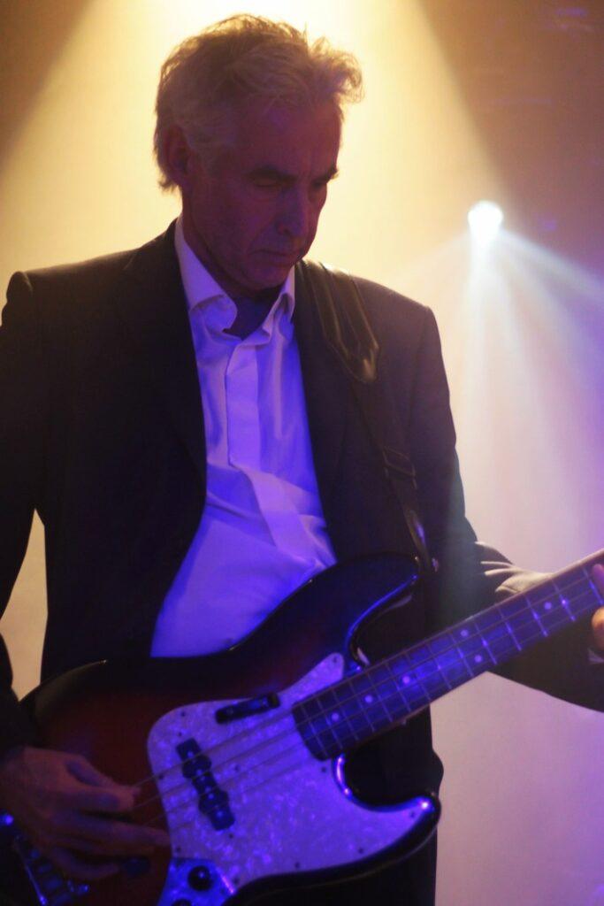 Bassiste élégant costume noir chemise blanche basse Jazz manche violet éclairage oranger fumée scène concert