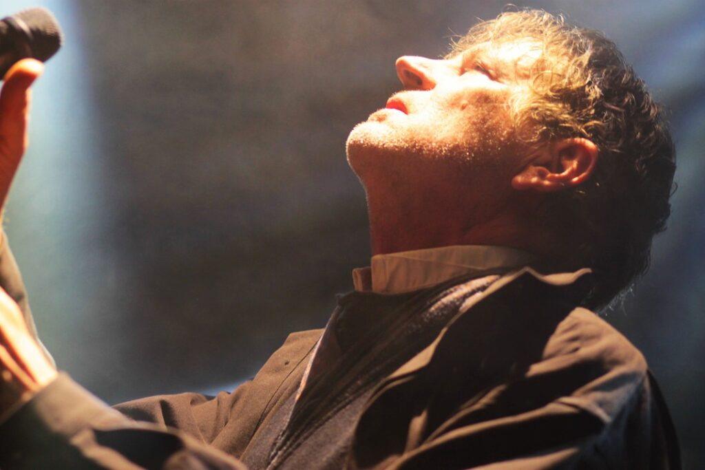 Chanteur homme aux cheveux bouclés en costume sombre et chemise blanche, chantant passionnément au micro sur scène, éclairage contre-jour dramatique ambiance concert live.