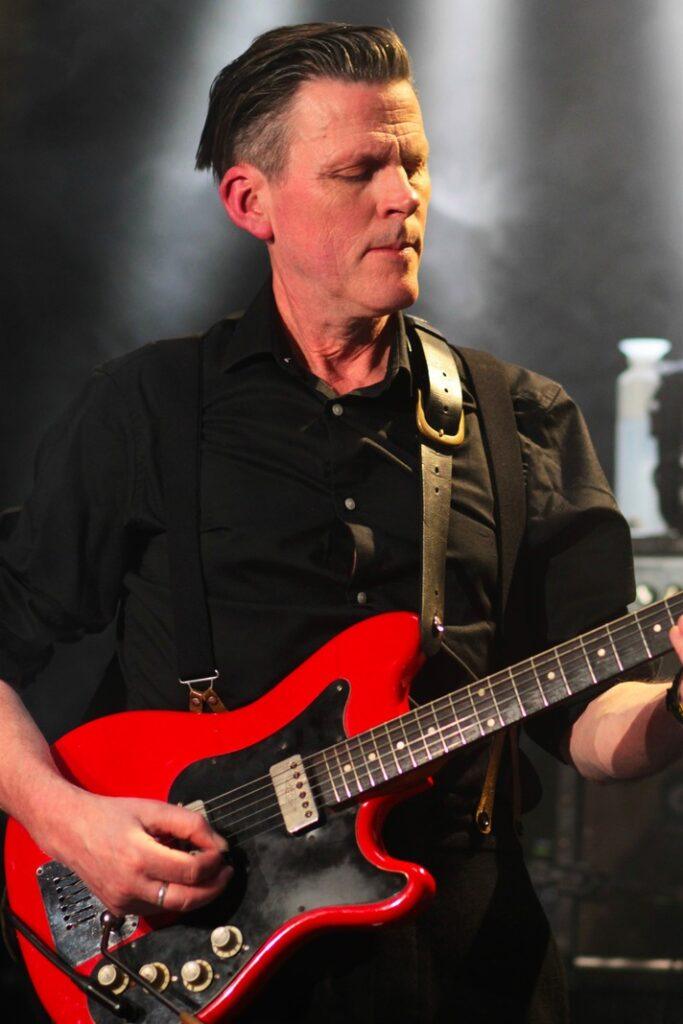 Guitariste rock homme aux cheveux gominés rasés côtés, jouant guitare électrique rouge sur scène concert, chemise noire bretelles cuir, éclairage spot ambiance live.