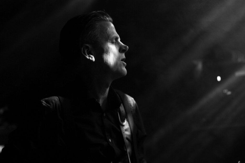 Portrait profil en noir et blanc d'un musicien aux cheveux gominés rasés côtés, regard intense éclairé en contre-jour sur scène de concert, ambiance dramatique live.