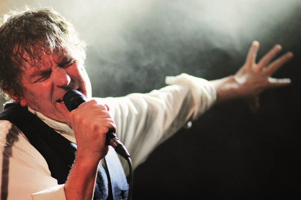 Chanteur énergique sur scène de concert, cheveux ébouriffés, chemise blanche gilet noir au micro, bras tendu passionné, éclairage brumeux ambiance live intense.