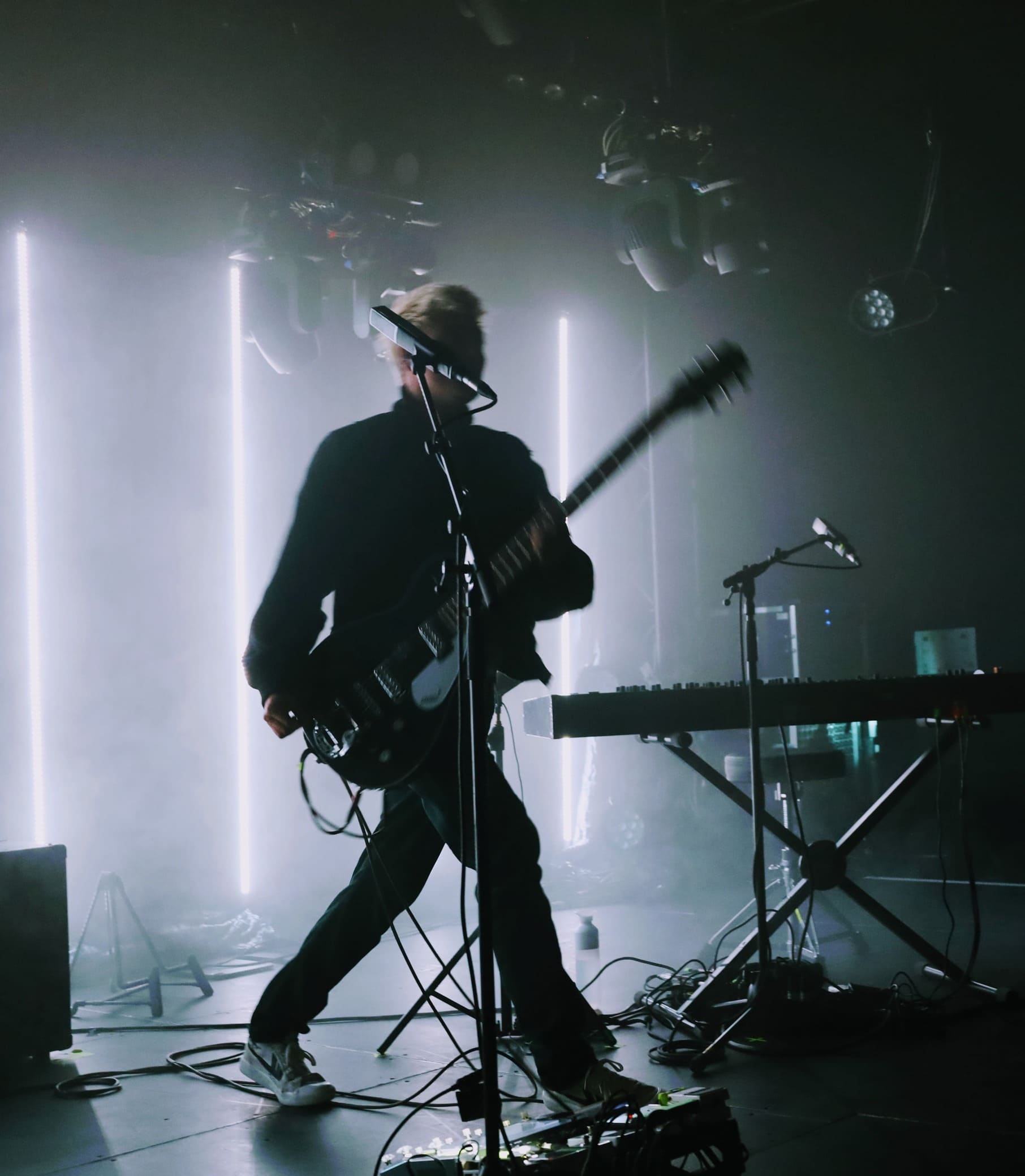 Guitariste blond mouvement guitare noire électrique synthé néons verticaux fumée scène concert sombre