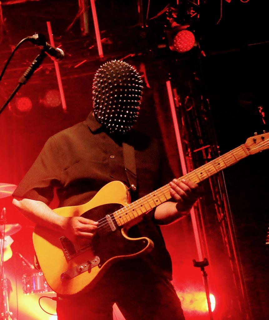 Gros plan du guitariste masqué de Skip the Use jouant de sa guitare électrique de style Telecaster sous une lumière rouge saturée.