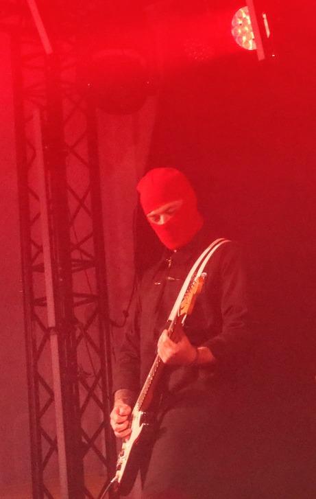 Le guitariste de Skip the Use jouant sur scène avec un masque de ski rouge et une combinaison noire sous des lumières rouges saturées.