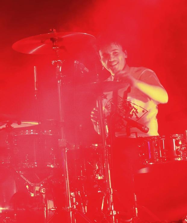 Le batteur de Skip the Use souriant derrière sa batterie en plein concert, éclairé par une lumière rouge intense.
