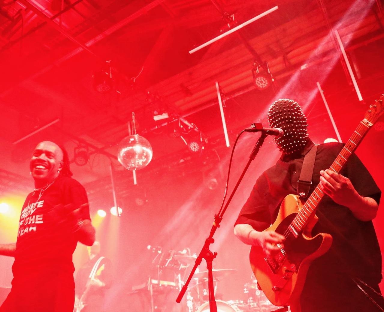 Mat Bastard souriant aux côtés du guitariste masqué de Skip the Use sur une scène de concert éclairée en rouge avec une boule à facettes.