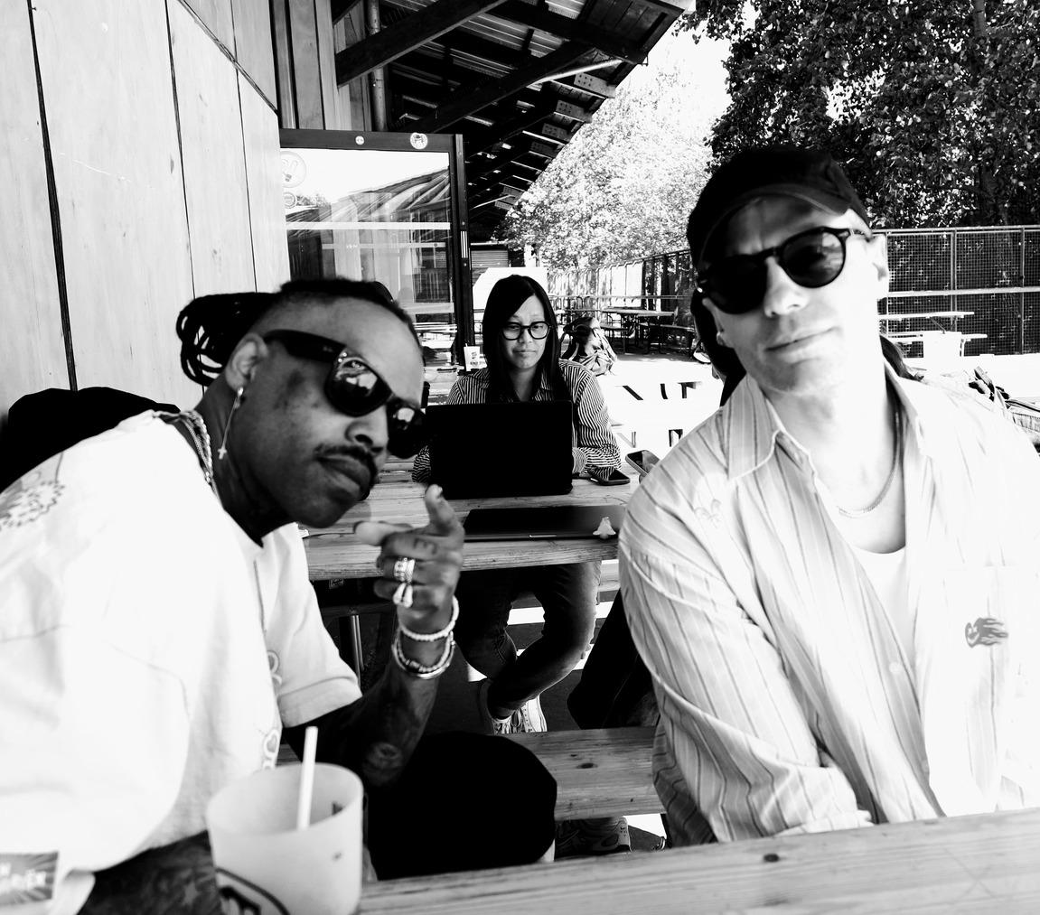 Photo en noir et blanc de Mat Bastard et d'un membre de Skip the Use assis en terrasse, avec une personne travaillant sur un ordinateur en arrière-plan.