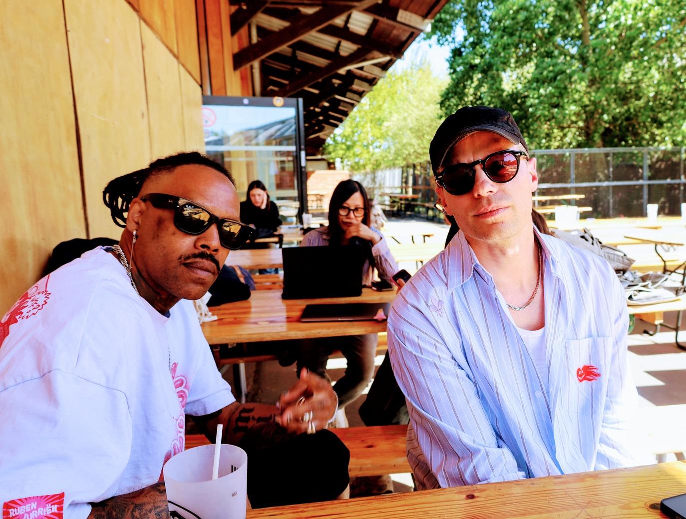 Photo de Mat Bastard et d'un membre de Skip the Use assis en terrasse, avec une personne travaillant sur un ordinateur en arrière-plan.