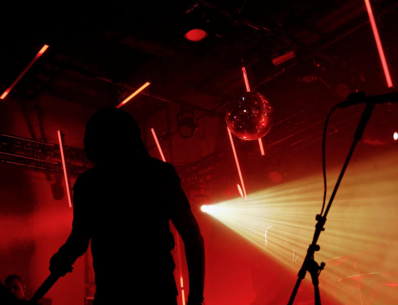 Vue de dos d'un artiste sur scène dans une ambiance de concert rock avec éclairages néons rouges et faisceaux lumineux blancs.
