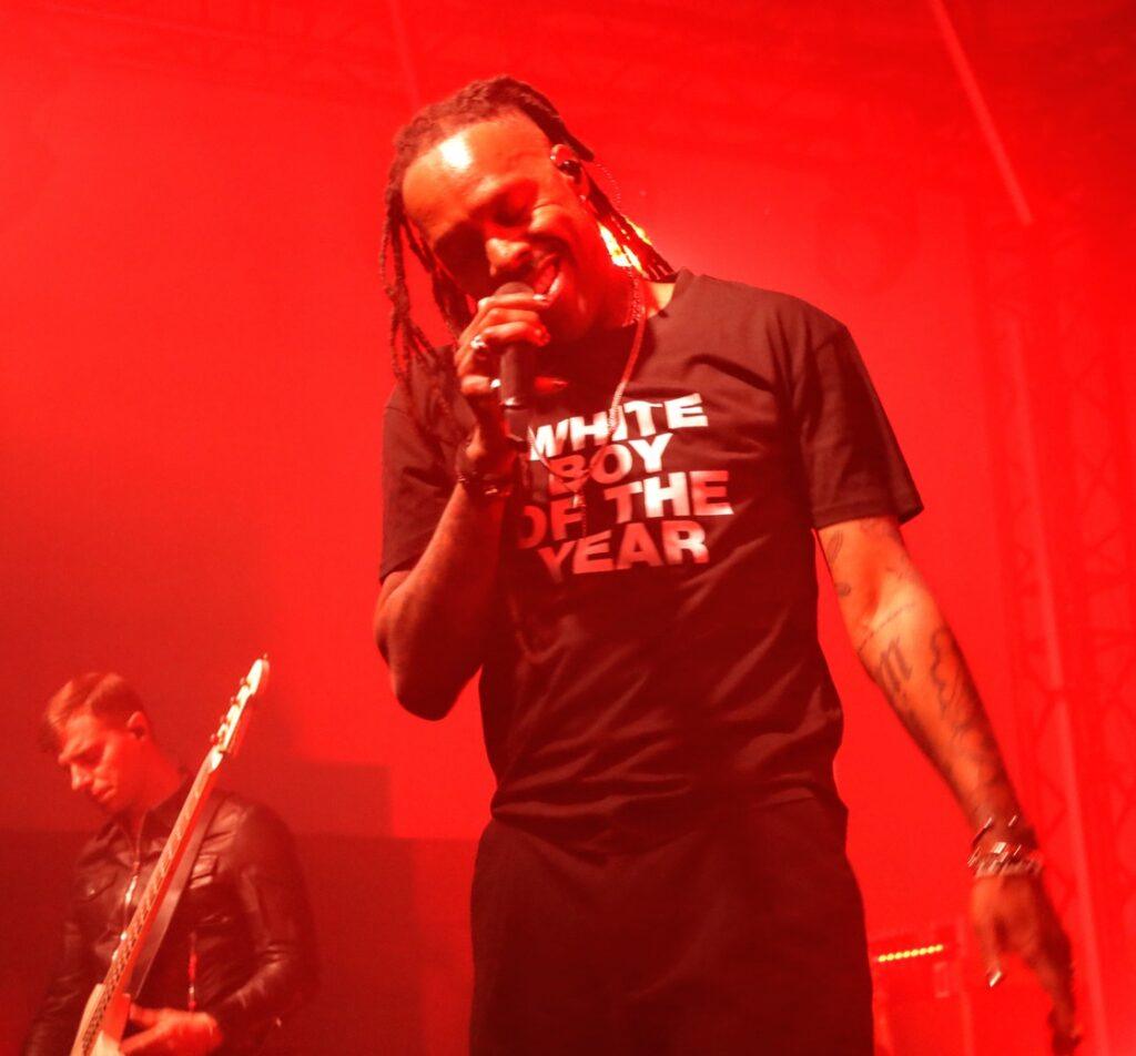 Mat Bastard, le chanteur de Skip the Use, souriant au micro sur scène avec un t-shirt "White Boy of the Year" sous des lumières rouges.