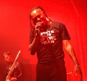 Mat Bastard, le chanteur de Skip the Use, souriant au micro sur scène avec un t-shirt "White Boy of the Year" sous des lumières rouges.