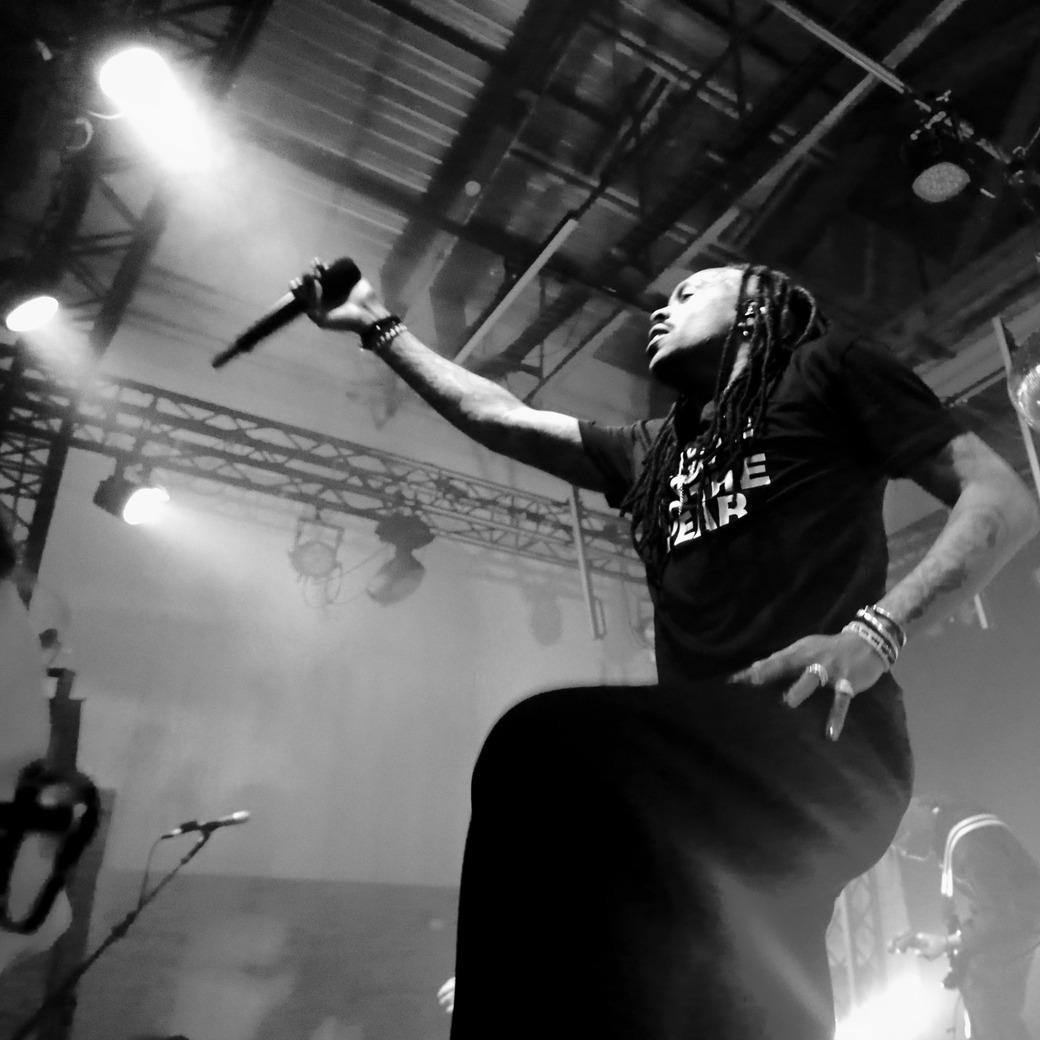 Vue en contre-plongée de Mat Bastard (Skip the Use) sur scène, micro à la main et bras tendu, dans une ambiance de concert rock en noir et blanc.