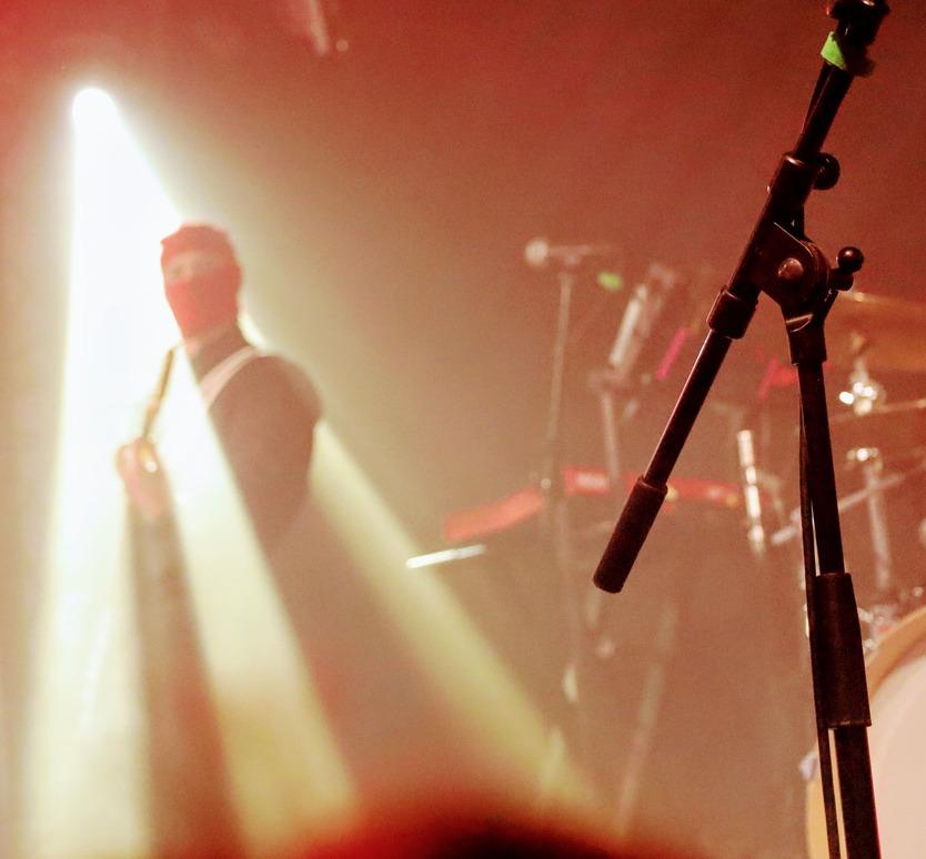 Vue de la scène de Skip the Use avec un pied de micro au premier plan et le guitariste masqué en arrière-plan sous un faisceau de lumière blanche.