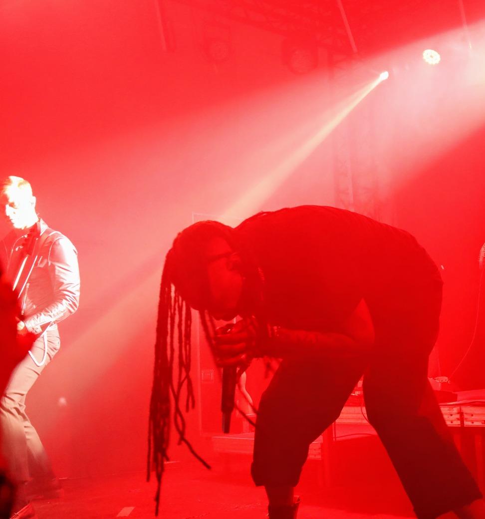 Chanteur de Skip the Use, penché en avant sur scène avec son micro, éclairé par une lumière rouge intense et un projecteur blanc.