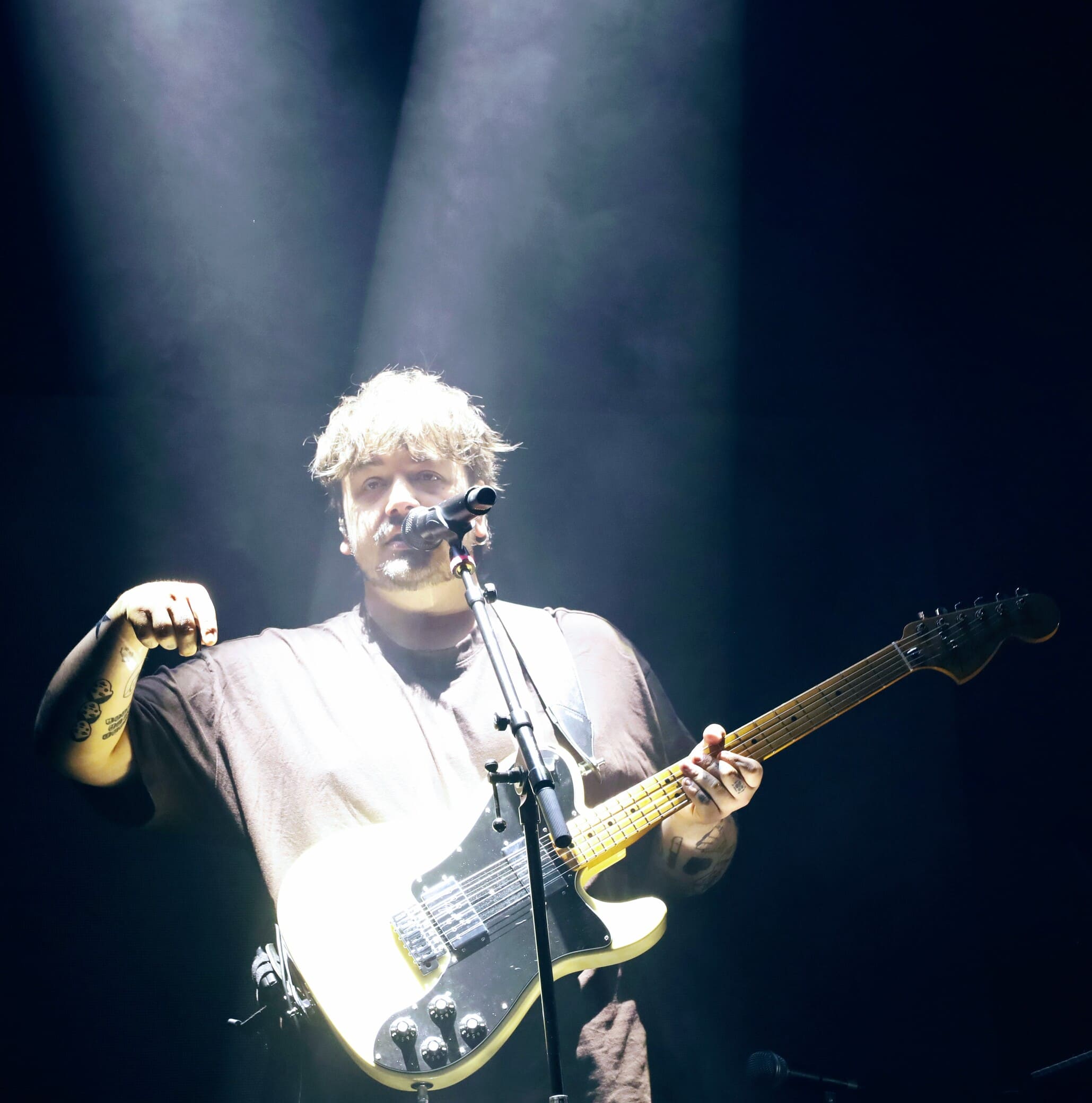 L'artiste Stgraal sur scène sous un spot lumineux, une main levée, chantant dans un micro et tenant sa guitare électrique.