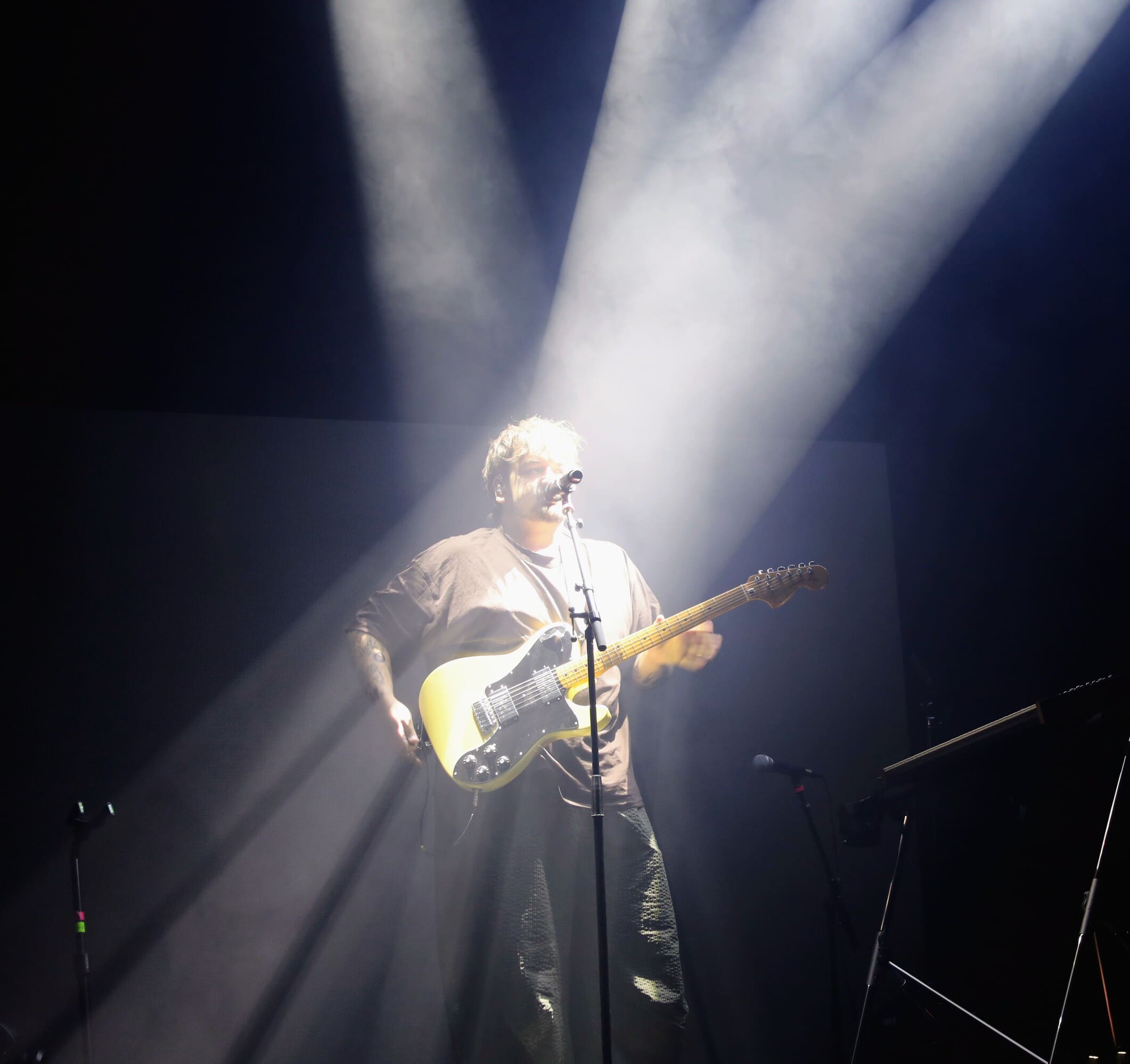 L'artiste Stgraal sur scène au milieu de faisceaux de lumière blanche, jouant de la guitare électrique et chantant face à son micro.