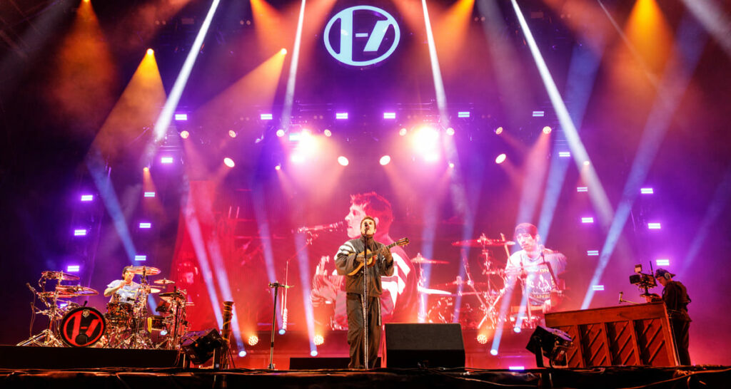 Concert de Twenty One Pilots sur la grande scène éclairée par des projecteurs violets et oranges avec logo suspendu.