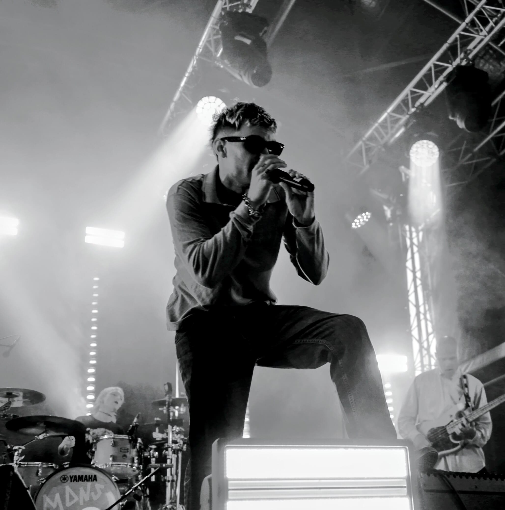 Chanteur du groupe MDNS en pleine performance sur scène, micro à la main, devant le batteur et le bassiste sous les projecteurs en noir et blanc.