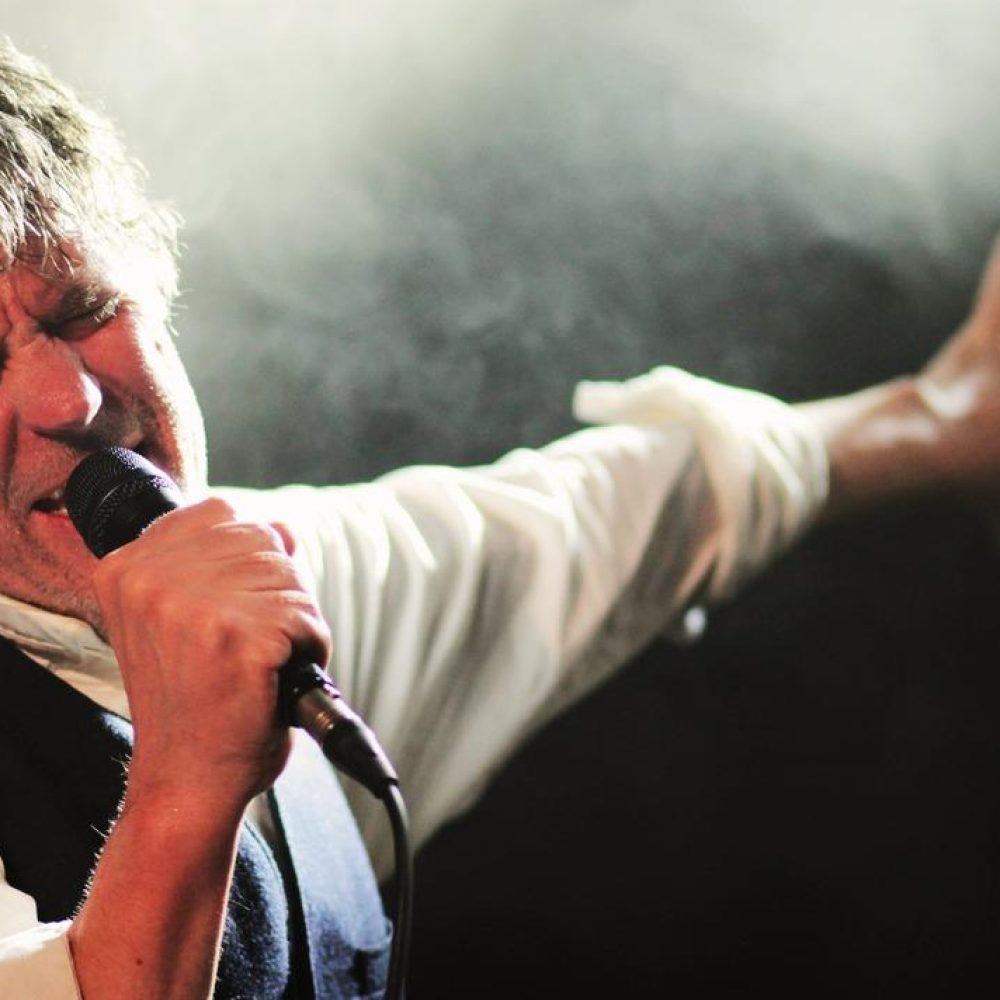 Chanteur énergique sur scène de concert, cheveux ébouriffés, chemise blanche gilet noir au micro, bras tendu passionné, éclairage brumeux ambiance live intense.
