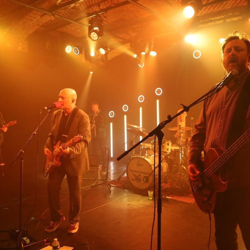 Groupe de rock sur scène jaune-orangée : chanteur barbu au micro central, guitariste chauve à gauche, bassiste barbu à droite jouant basse, batterie, éclairages spots ronds et bâtons lumineux, ambiance live chaleureuse.
